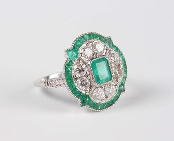 A platinum, emerald and diamond ring in an Art Deco inspired cluster design, mounted with the cut cornered rectangular step cut emerald within a quatr